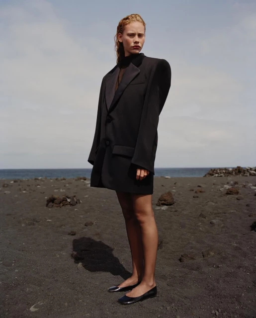 A fair-skinned woman in an oversized black blazer and black flats stands on a dark sand beach under a cloudy blue sky by the ocean.