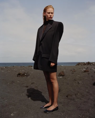 A fair-skinned woman in an oversized black blazer and black flats stands on a dark sand beach under a cloudy blue sky by the ocean.