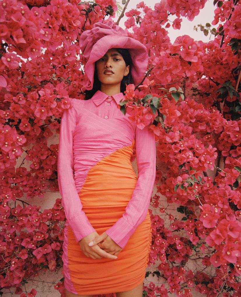 Woman poses in a pink and orange dress with matching hat, surrounded by vibrant pink flowers, creating a striking color contrast.