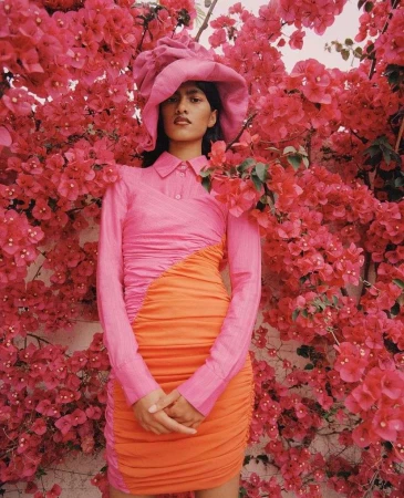 Woman poses in a pink and orange dress with matching hat, surrounded by vibrant pink flowers, creating a striking color contrast.