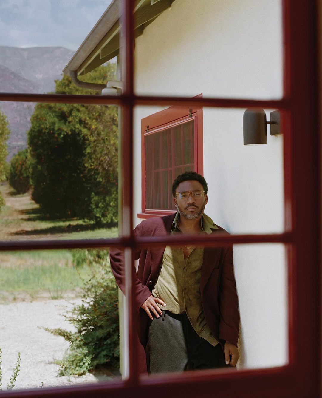 Adult male poses in front of a window with a burgundy frame, wearing a burgundy jacket, light green shirt, and dark pants, outdoor view.