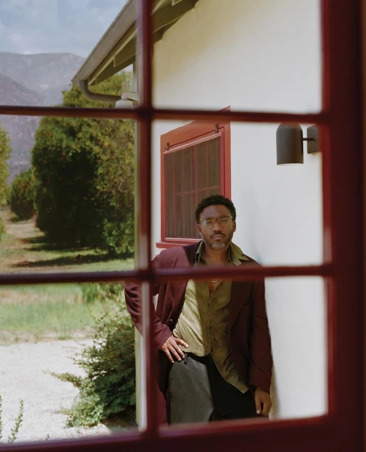 Adult male poses in front of a window with a burgundy frame, wearing a burgundy jacket, light green shirt, and dark pants, outdoor view.