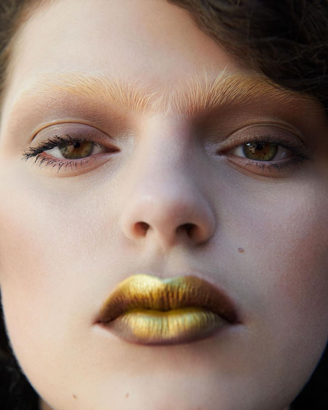 Close-up portrait of a young adult female with striking gold lipstick, light eyebrows, and brown eyes. Beauty shot with natural lighting.