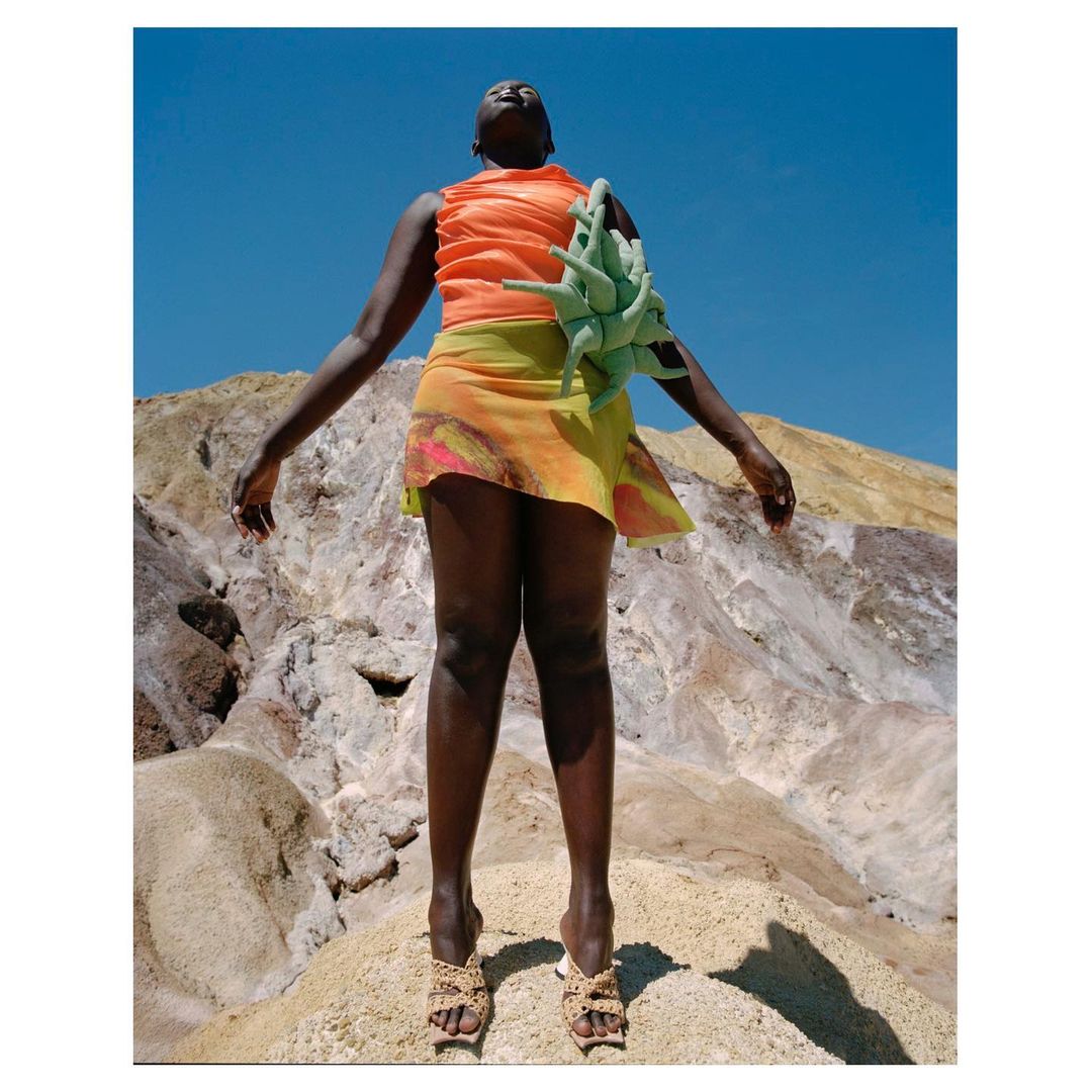 Black woman in orange top and patterned skirt with sculptural green accessory standing on a rocky outcrop under a clear blue sky