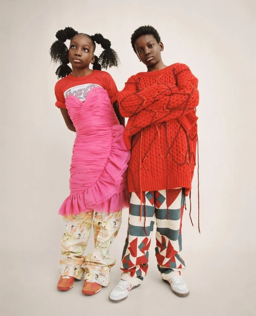 Two young fashion models pose in a photo studio. One wears a red sweater and patterned pants, and the other wears a pink dress and patterned pants.