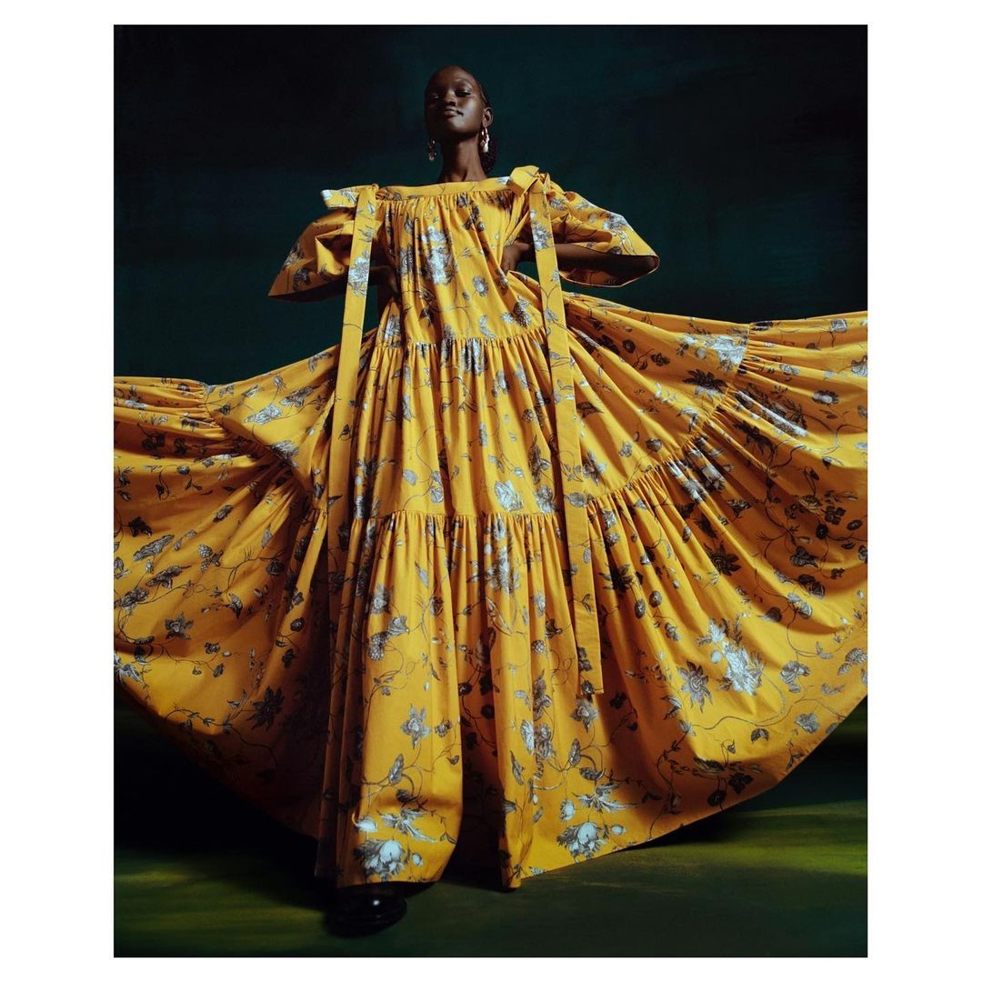Dark-skinned woman in long yellow dress with floral pattern posing in a studio with dark green background; she's wearing dangling earrings.
