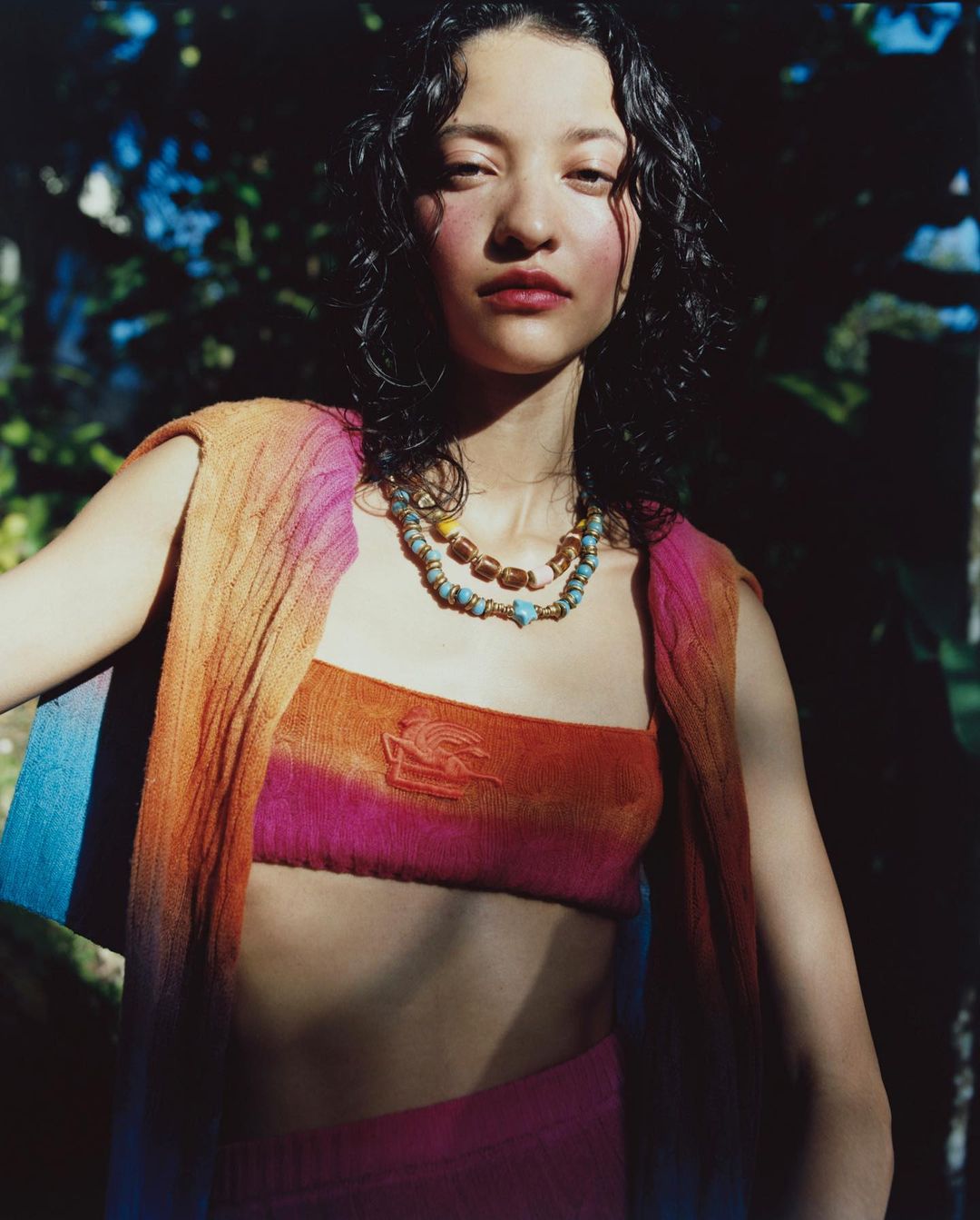 Young woman poses outdoors in a colorful knit top and skirt with beaded necklaces, soft lighting, and a blurry foliage background.