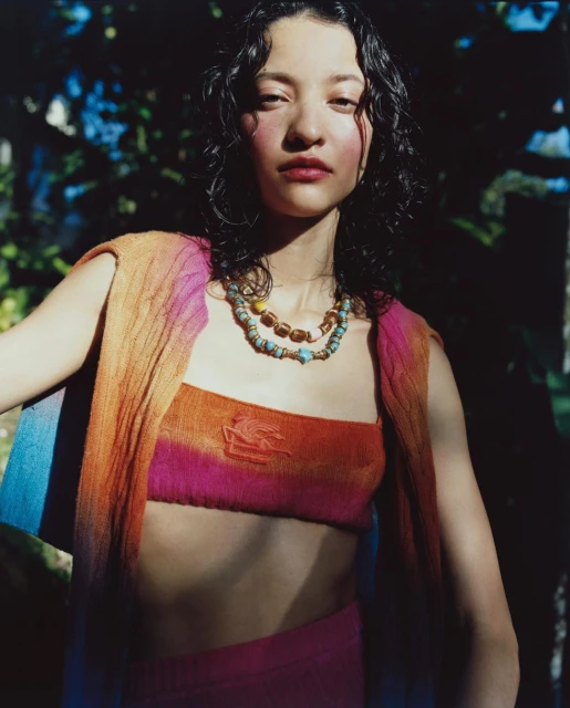 Young woman poses outdoors in a colorful knit top and skirt with beaded necklaces, soft lighting, and a blurry foliage background.