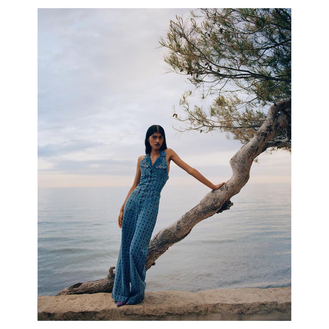 A woman in a denim patterned jumpsuit stands on a ledge near the ocean, leaning against a tree in a fashion style editorial shot.