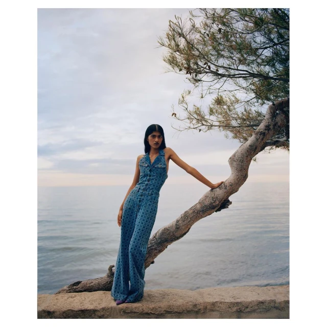 A woman in a denim patterned jumpsuit stands on a ledge near the ocean, leaning against a tree in a fashion style editorial shot.