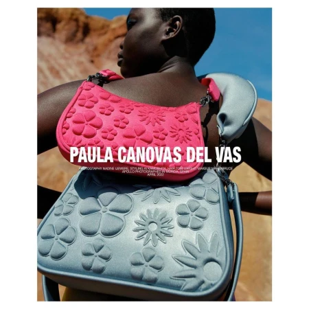 Black woman wearing two stacked flower-embossed shoulder bags in pink and silver against a rocky landscape and blue sky.
