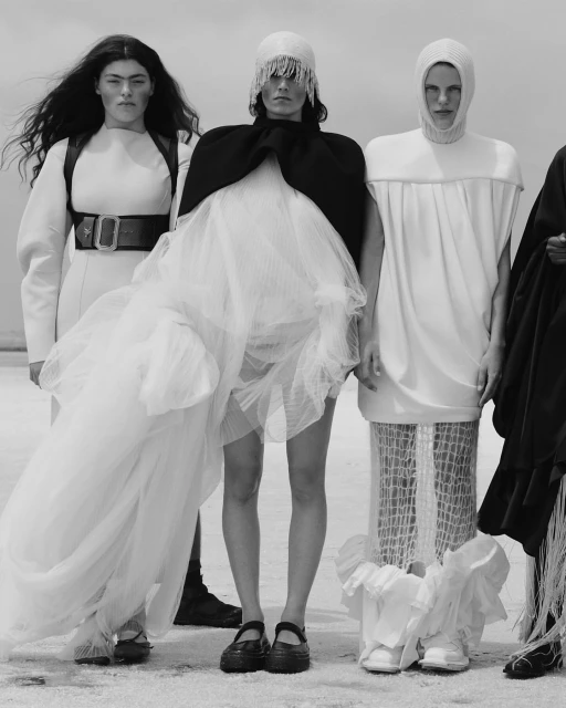 Black-and-white fashion photo of five female models wearing uniquely designed gowns and dresses on a sandy, bright, outdoor setting.