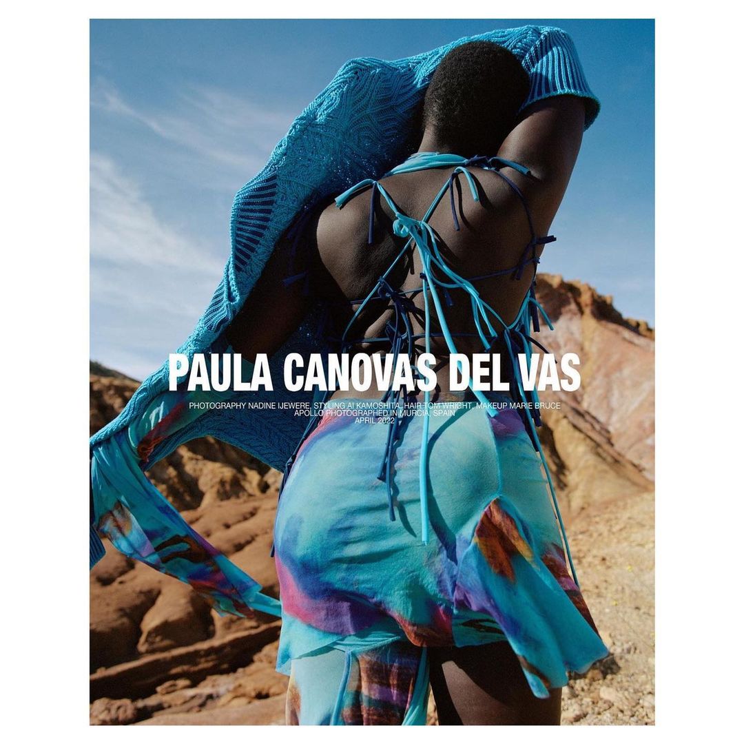 Fashion editorial shot of an African female model in a blue patterned dress and shawl posing outdoors in front of a desert landscape.
