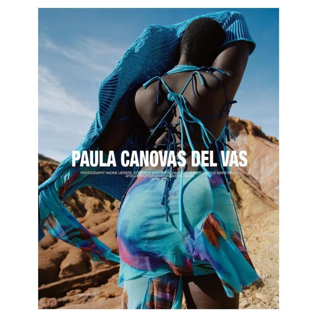 Fashion editorial shot of an African female model in a blue patterned dress and shawl posing outdoors in front of a desert landscape.