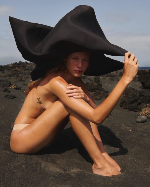 A single nude female model sits on a black sandy beach wearing a large black hat, posed with arms around her legs and ocean in the background.