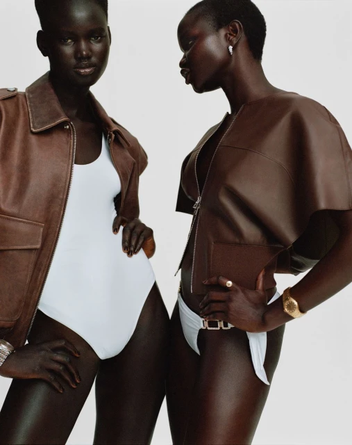 Two African-descent women pose in white bodysuits and brown leather jackets against a bright backdrop, showcasing fashion and style.