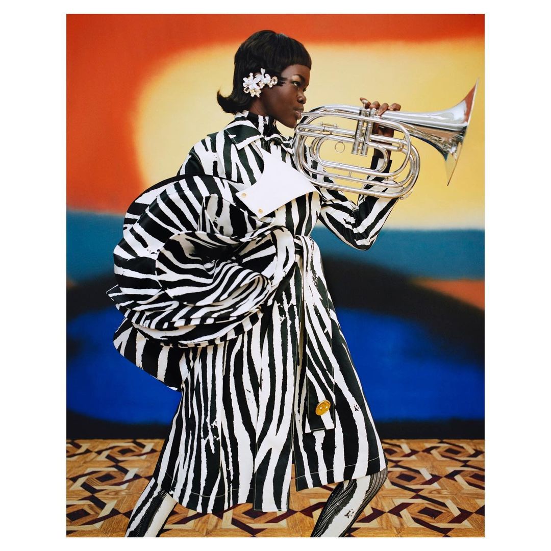 A dark-skinned woman in a zebra-striped outfit plays a silver French horn against a colorful backdrop and geometric floor pattern. 