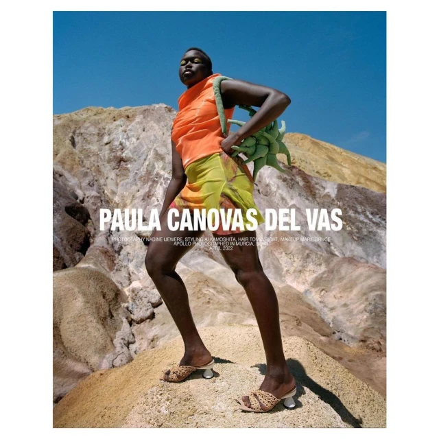 A fashion shot of a Black woman in an orange top and yellow skirt, holding a green bag, posing against a textured, rocky landscape under a blue sky.