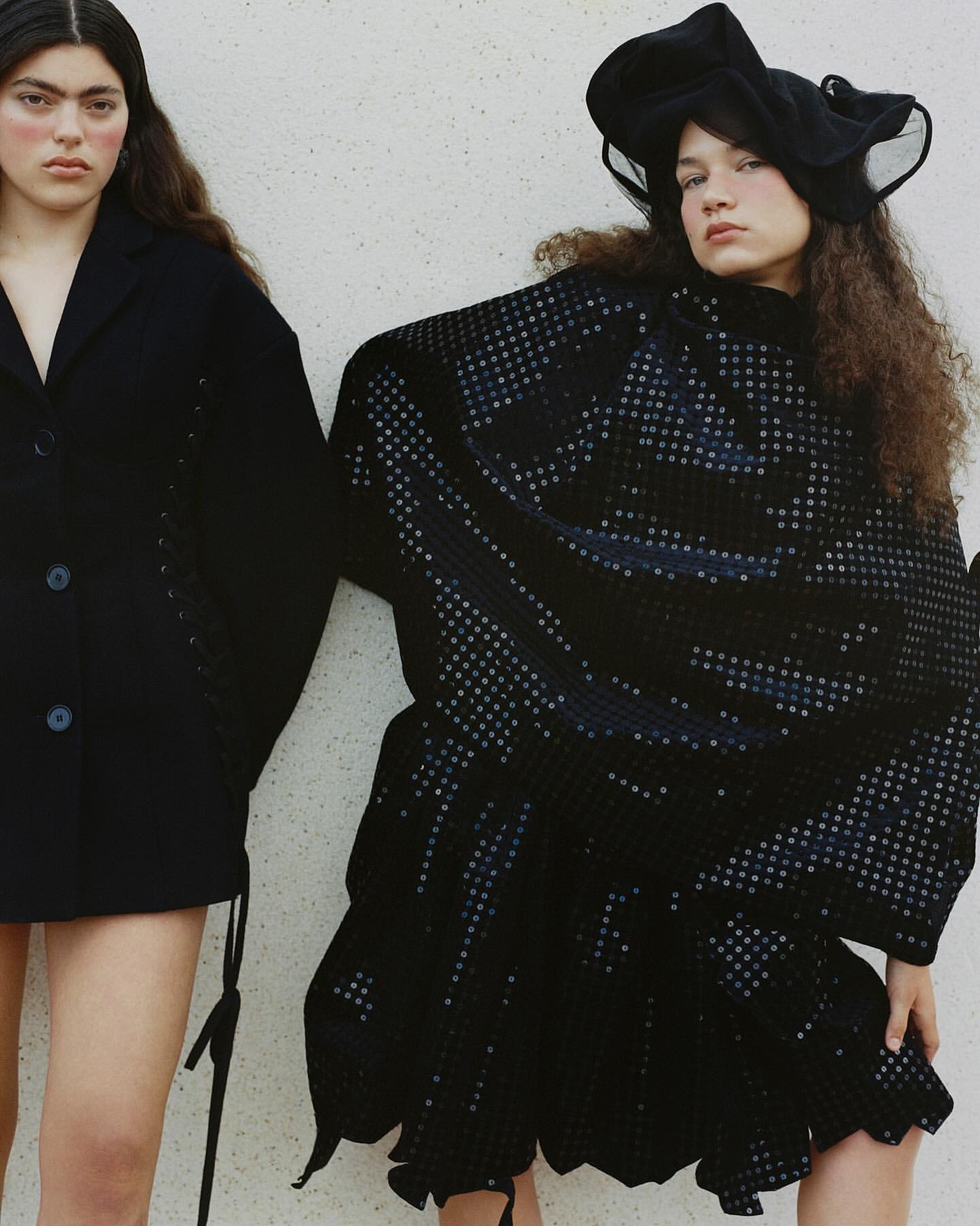Two women pose against a white wall wearing dark fashion; one in a blazer, the other in a dotted dress and hat in editorial style.