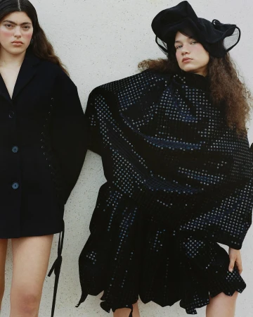 Two women pose against a white wall wearing dark fashion; one in a blazer, the other in a dotted dress and hat in editorial style.