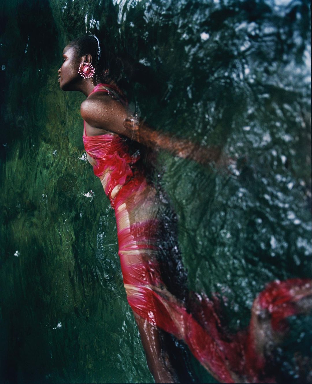 Woman in a pink dress swims underwater in a rocky river; she wears statement earrings and looks to the left in a fashion shot.