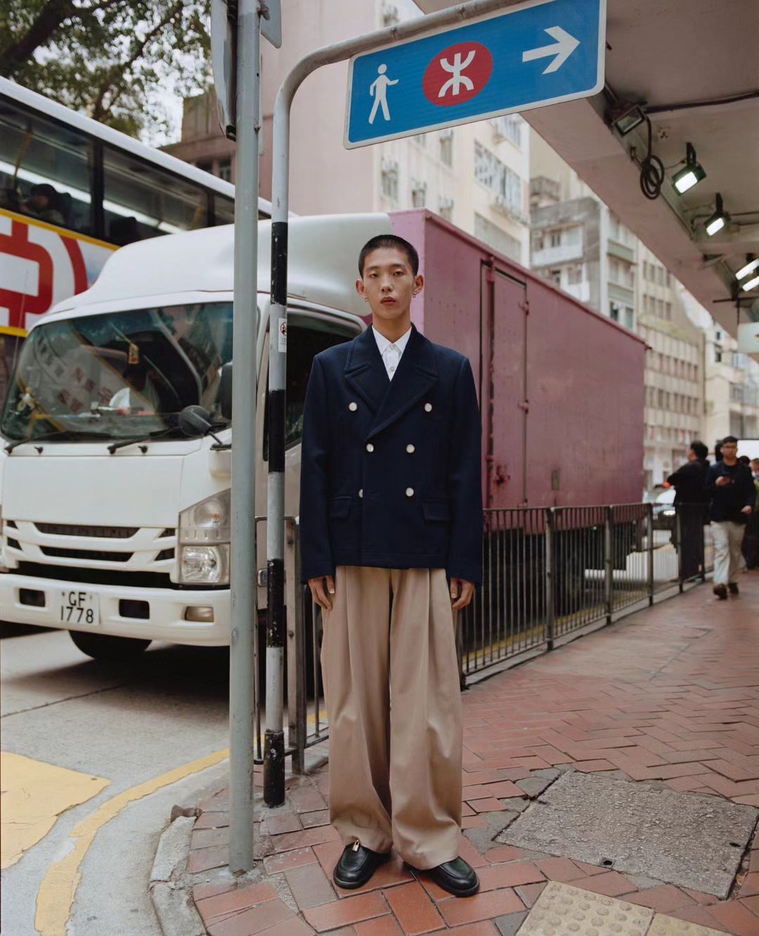 Adult male fashion model poses in a navy double-breasted blazer and wide-legged trousers on an urban sidewalk with city traffic.