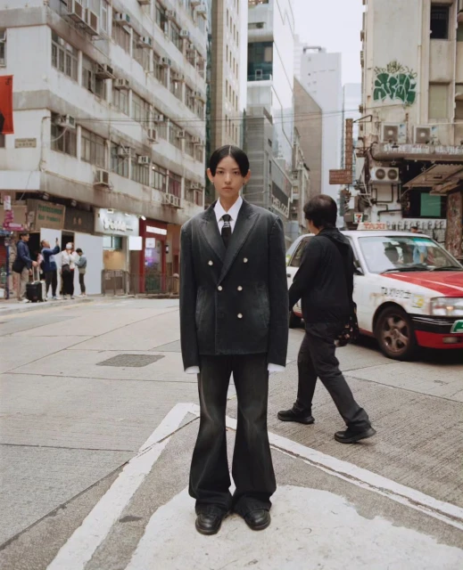 A person in a dark suit poses on a city street corner, next to a crosswalk with buildings and a taxi in the urban background.