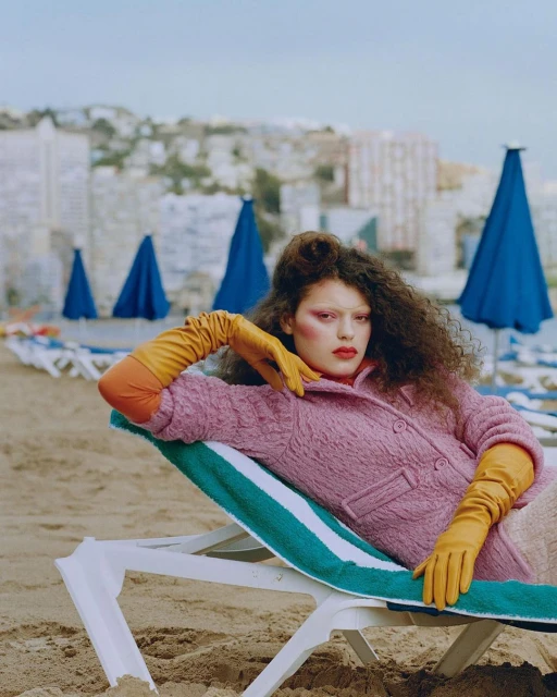 A woman in a pink coat and yellow gloves poses on a beach lounge chair against a backdrop of buildings and blue beach umbrellas.