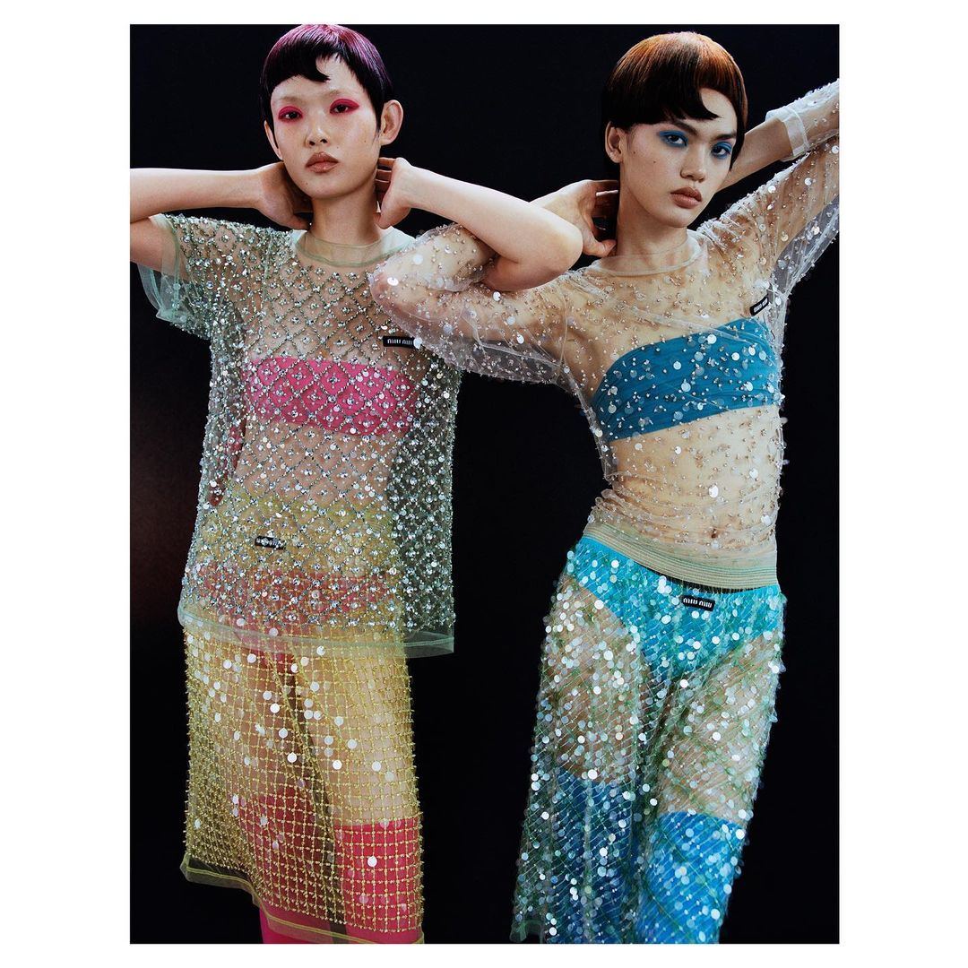 Two young Asian women in sheer, embellished dresses pose in front of a dark background. Fashion shot with trendy makeup and unique styling.