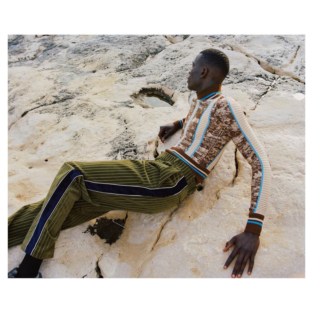 Dark-skinned male model poses on rocks wearing a patterned brown and tan sweater and olive pants with a blue stripe for fashion.