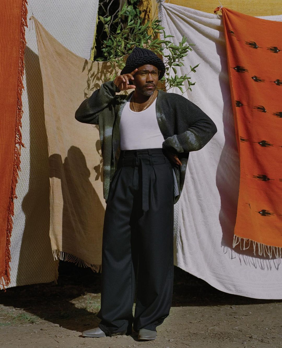 An adult male in a white top and loose black pants, wearing a dark knit hat and an open cardigan, stands outdoors in sunlight.