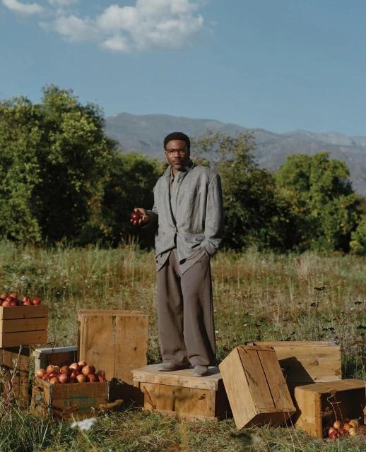 Adult man in stylish grey suit standing among wooden crates of apples in rural setting with mountain background under clear sky.