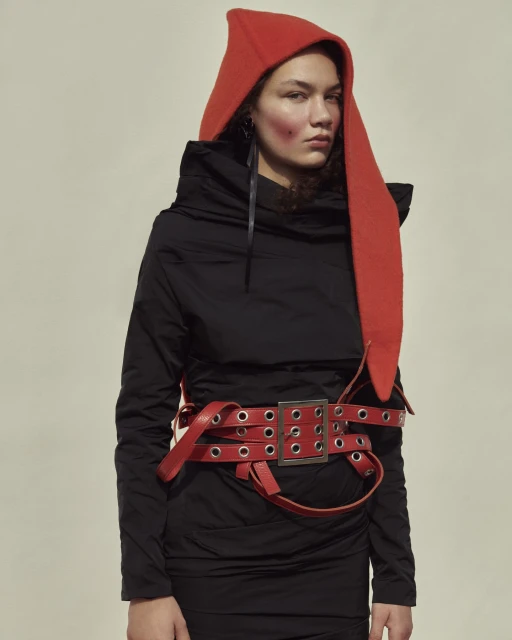 Woman in black dress with red hooded hat and red belt poses indoors with soft lighting for a fashion editorial shot.