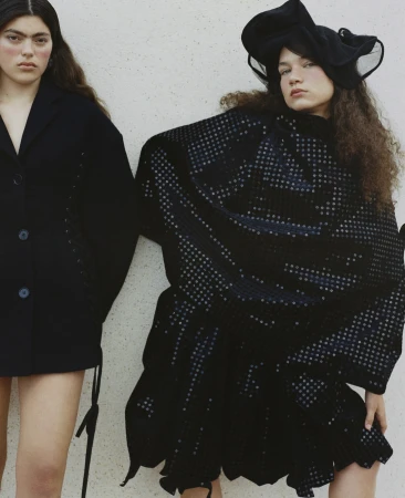 Two female models in avant-garde black attire against a plain textured wall photographed by Nadine Ijewere.