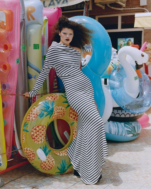 Model poses in a striped dress amidst colorful inflatable pool toys, capturing a vibrant summer aesthetic with a high-fashion edge.