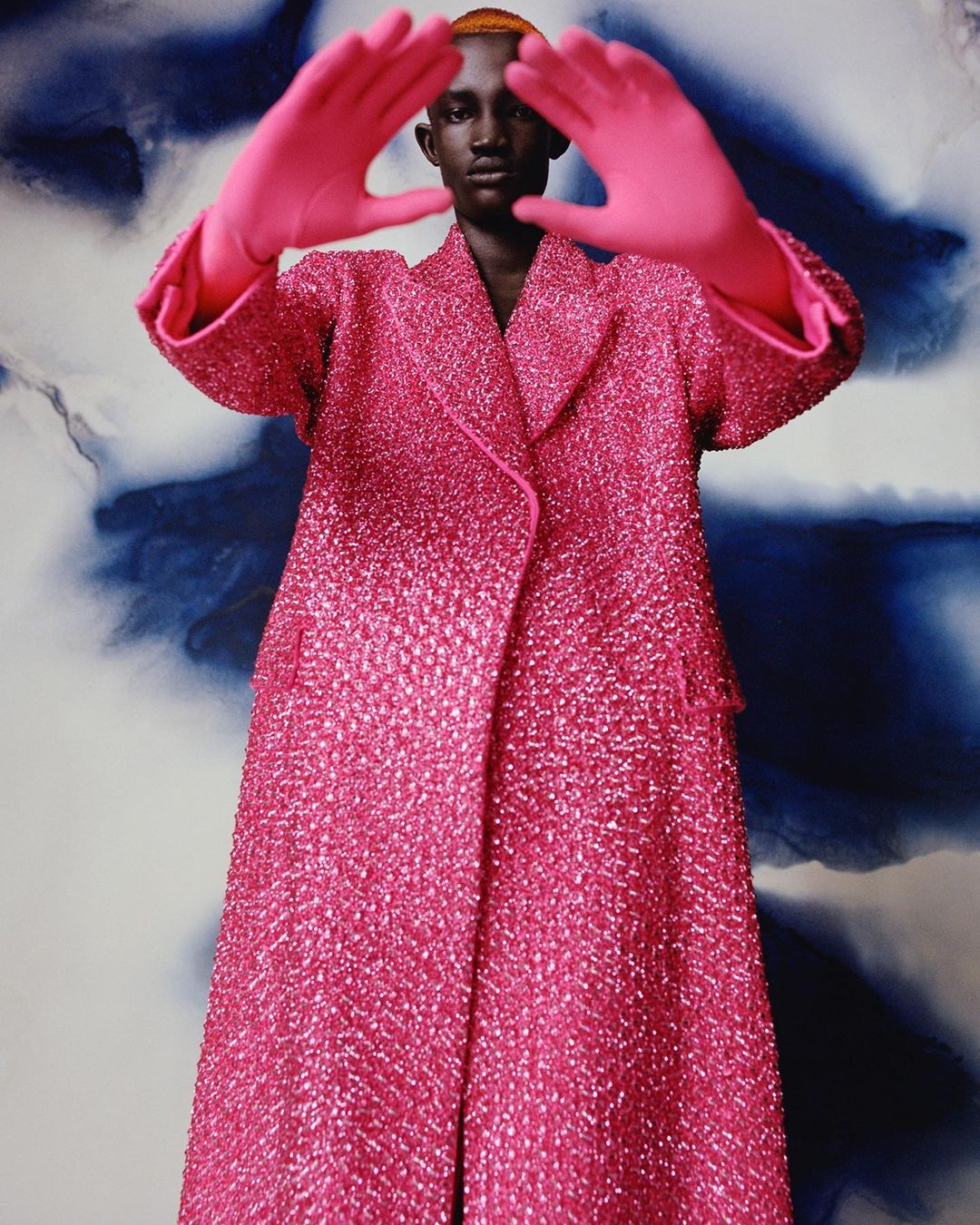 Fashion shot of a Black model in a pink coat and matching gloves framing their face against a blue and white patterned background.