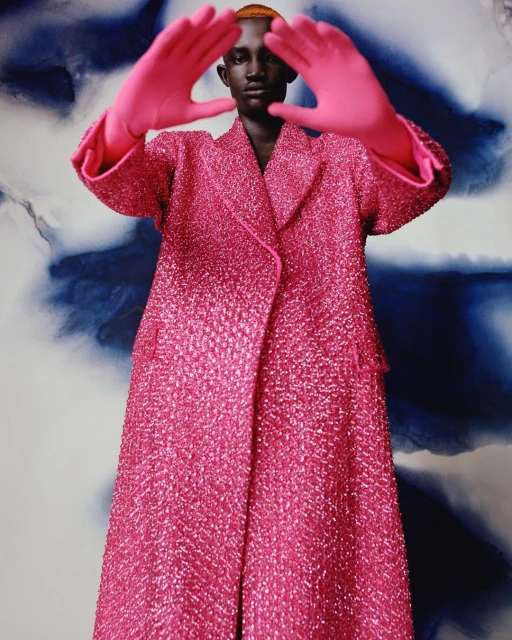 Fashion shot of a Black model in a pink coat and matching gloves framing their face against a blue and white patterned background.