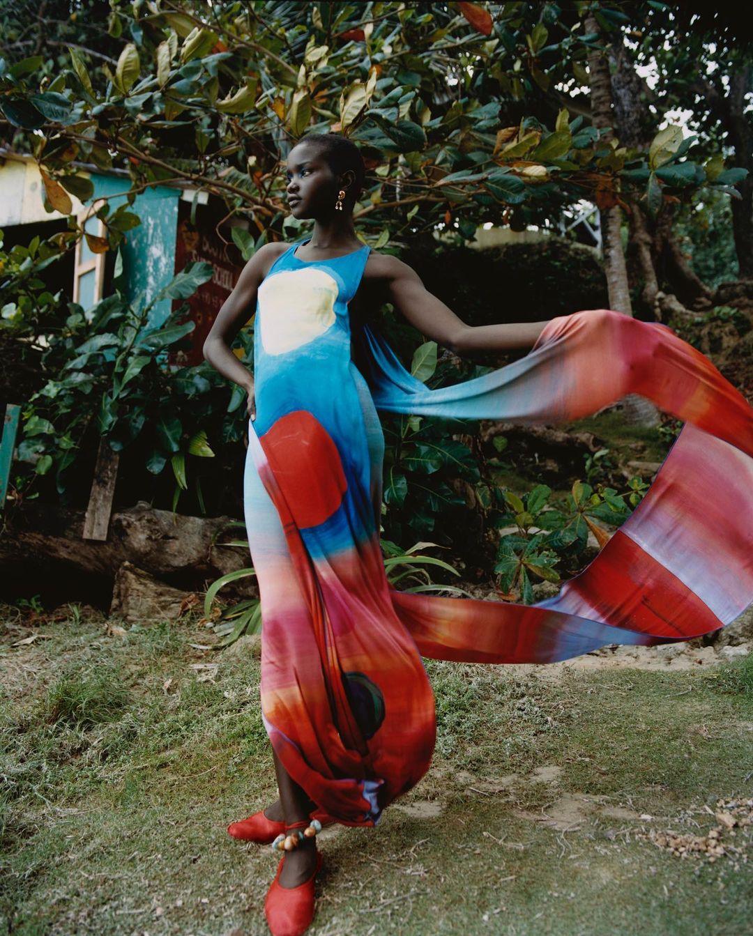 A stylish black woman in a flowing, colorful dress poses in a lush outdoor setting with a quaint building in the background.