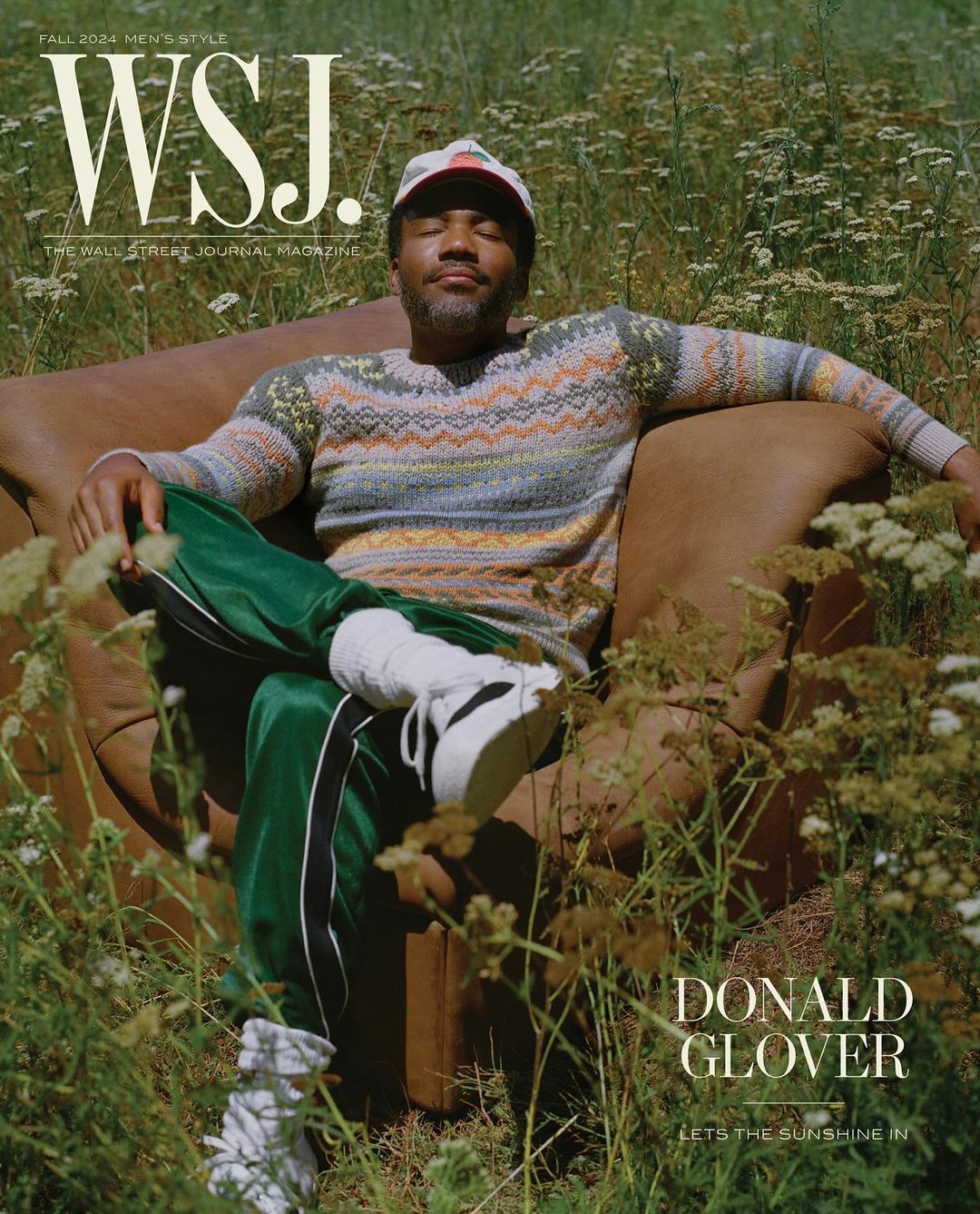 Donald Glover poses on brown leather armchair in a field of flowers for WSJ. Magazine, wearing a patterned sweater and green track pants.