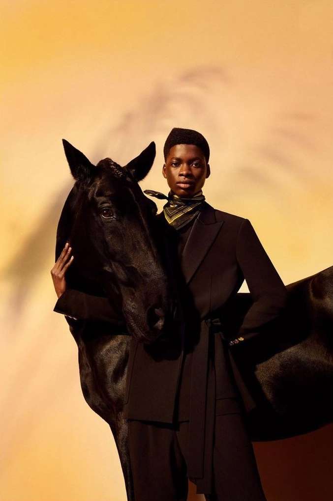 Fashion shot of an African American model in a black suit hugging a black horse in front of a warm, orange-toned background.