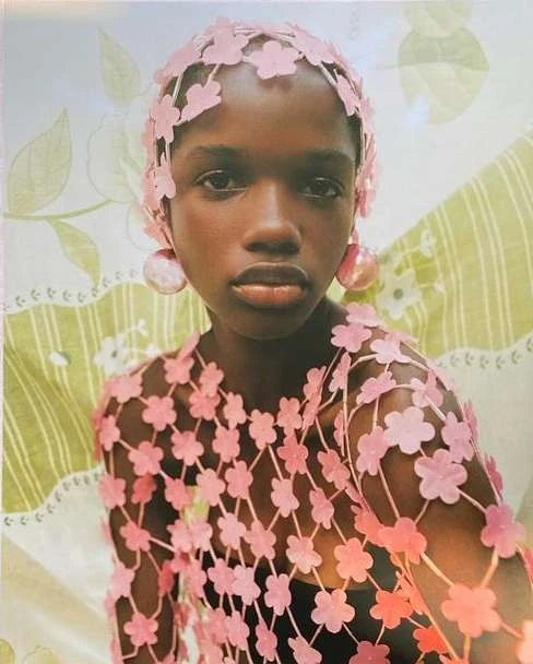 Headshot of a Black woman wearing a pink floral top and head covering, with matching earrings, set against a floral wallpaper backdrop.