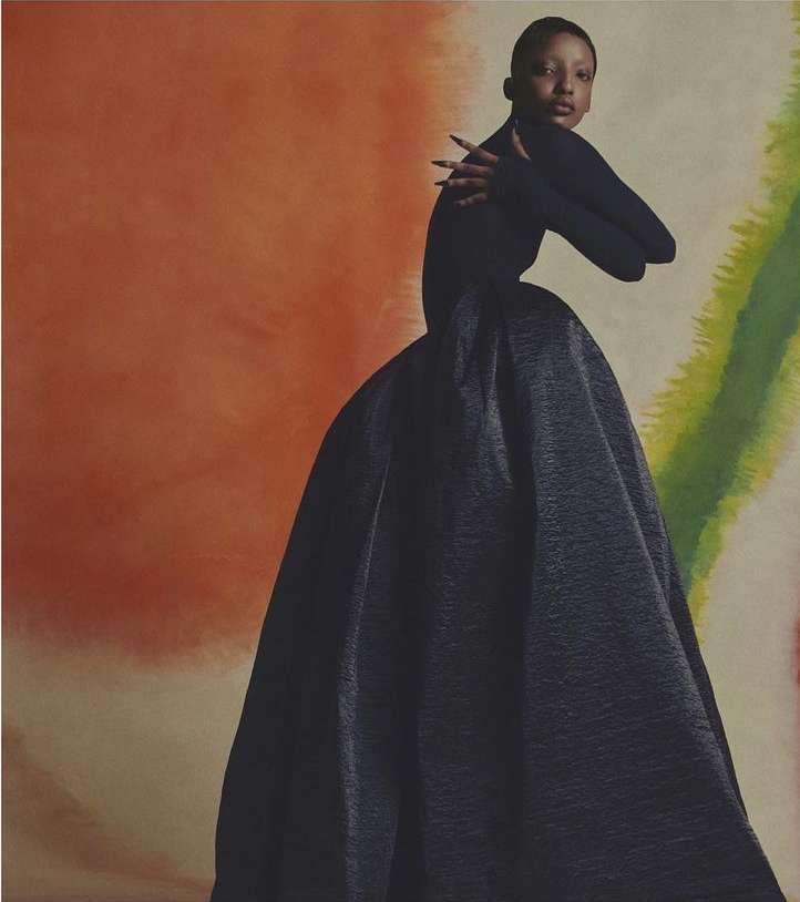 Adult female fashion model poses in black dress in front of an orange and beige painted background in a studio setting.
