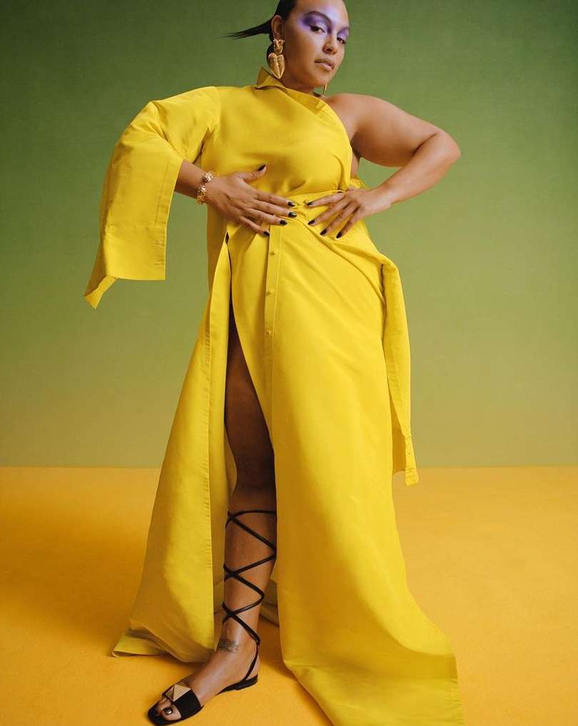 Woman in yellow asymmetrical dress, with cut-out shoulder and thigh-high split, accessorized with jewelry, stands in a studio with soft lighting.