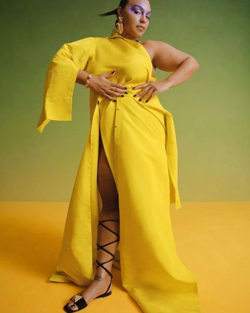 Woman in yellow asymmetrical dress, with cut-out shoulder and thigh-high split, accessorized with jewelry, stands in a studio with soft lighting.