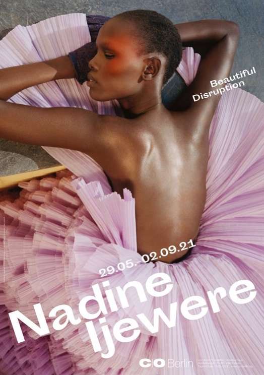 Dark-skinned woman poses in a light pink tulle dress. Shoulder-up shot; she rests her head on her arms looking to the left in a studio environment.