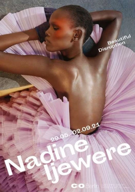 Dark-skinned woman poses in a light pink tulle dress. Shoulder-up shot; she rests her head on her arms looking to the left in a studio environment.