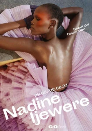 Dark-skinned woman poses in a light pink tulle dress. Shoulder-up shot; she rests her head on her arms looking to the left in a studio environment.