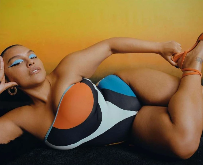 A stylish adult woman in a colorful swimsuit poses on a black fur-like surface in front of a blurred orange gradient background.