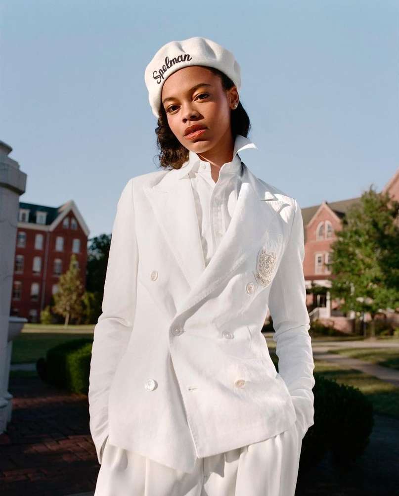 Elegant woman in a white suit and beret poses outdoors against a brick building. Spelman is embroidered on the beret.