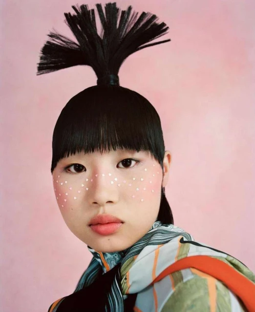 Close-up of an Asian female model with unique black pigtails, fair skin, and decorative heart-shaped makeup against a pink background.